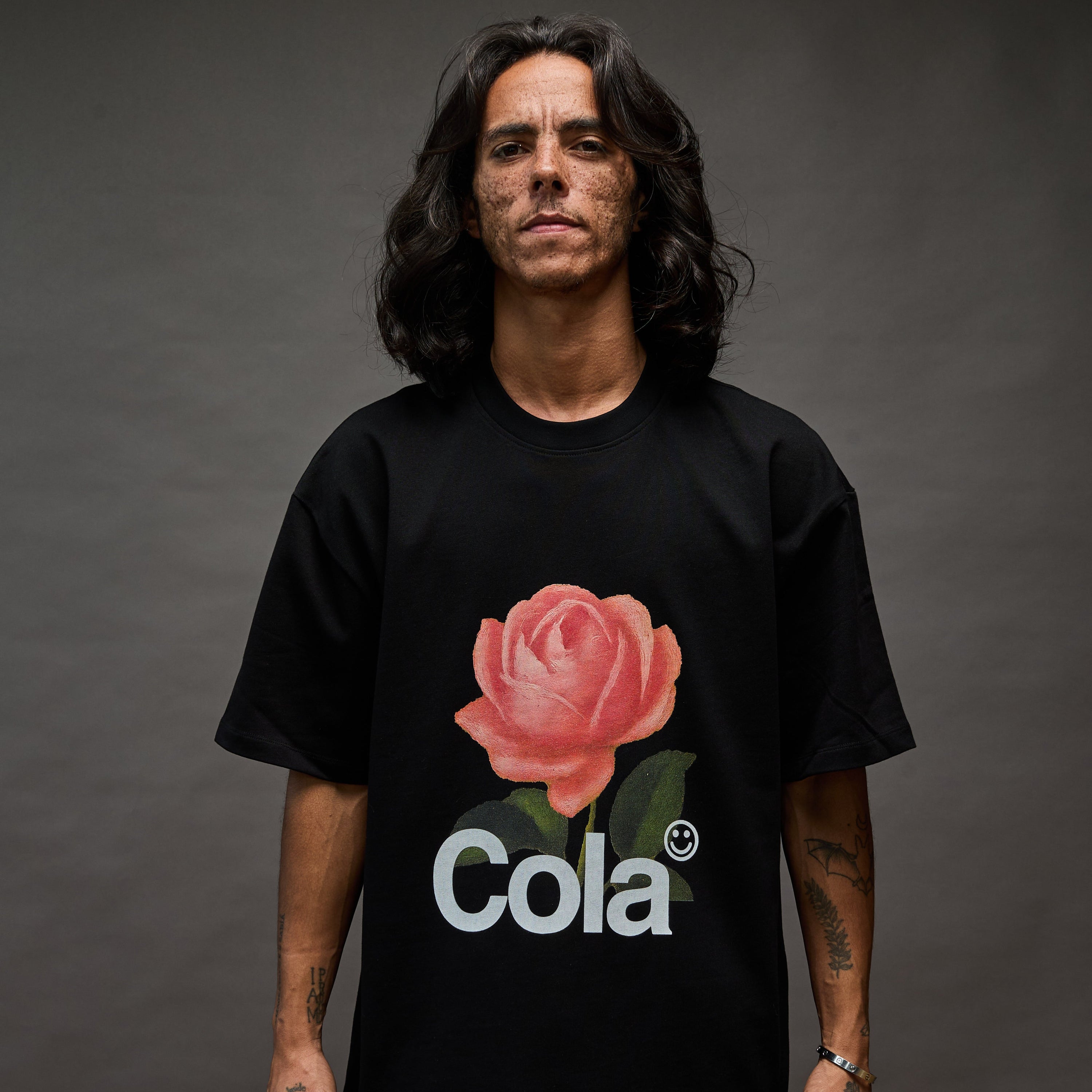 Person wearing a black t-shirt with a pink rose and 'Cola' text on a dark background