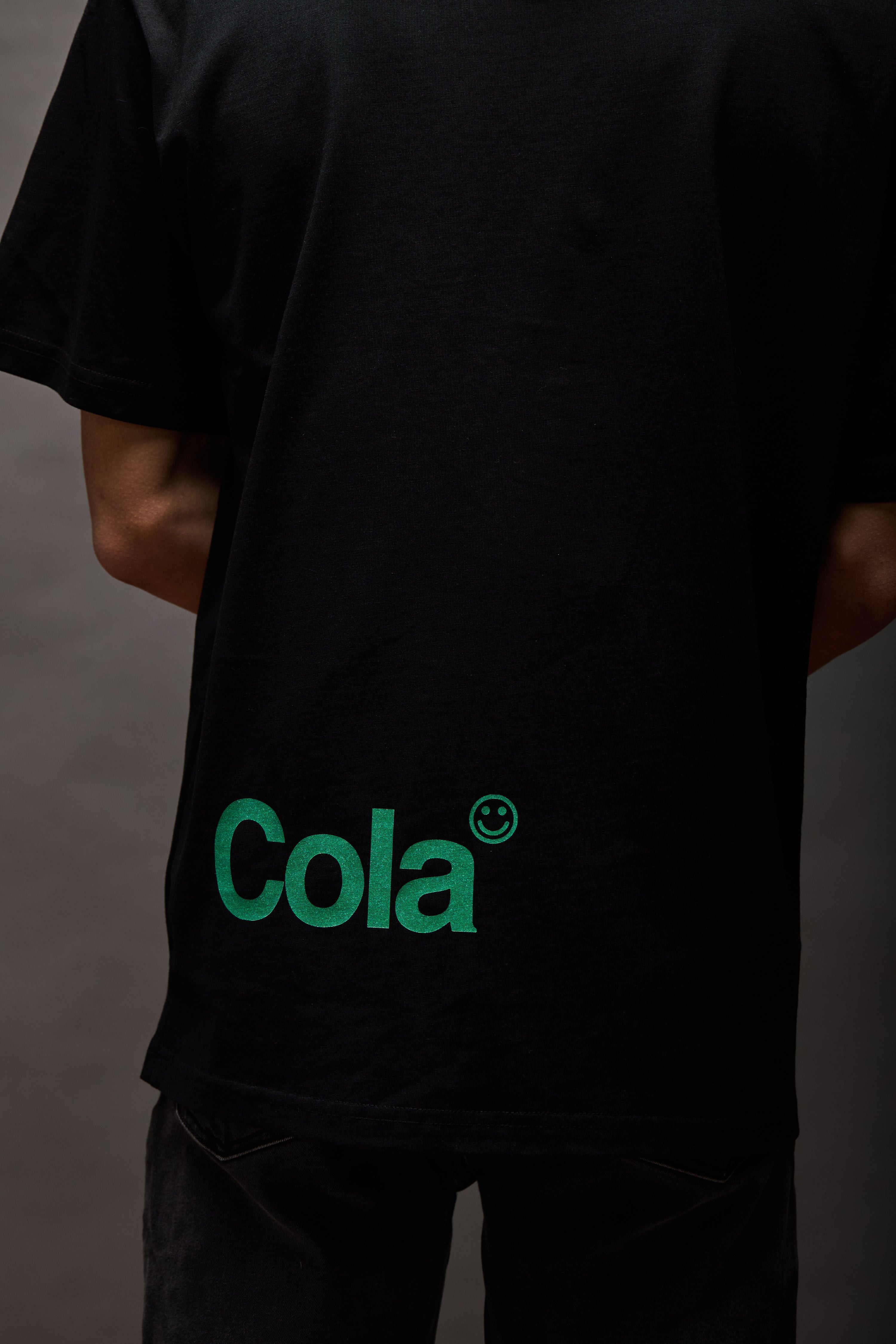 Back of black T-shirt with green “Cola” logo and small smiley mark printed near the lower back hem