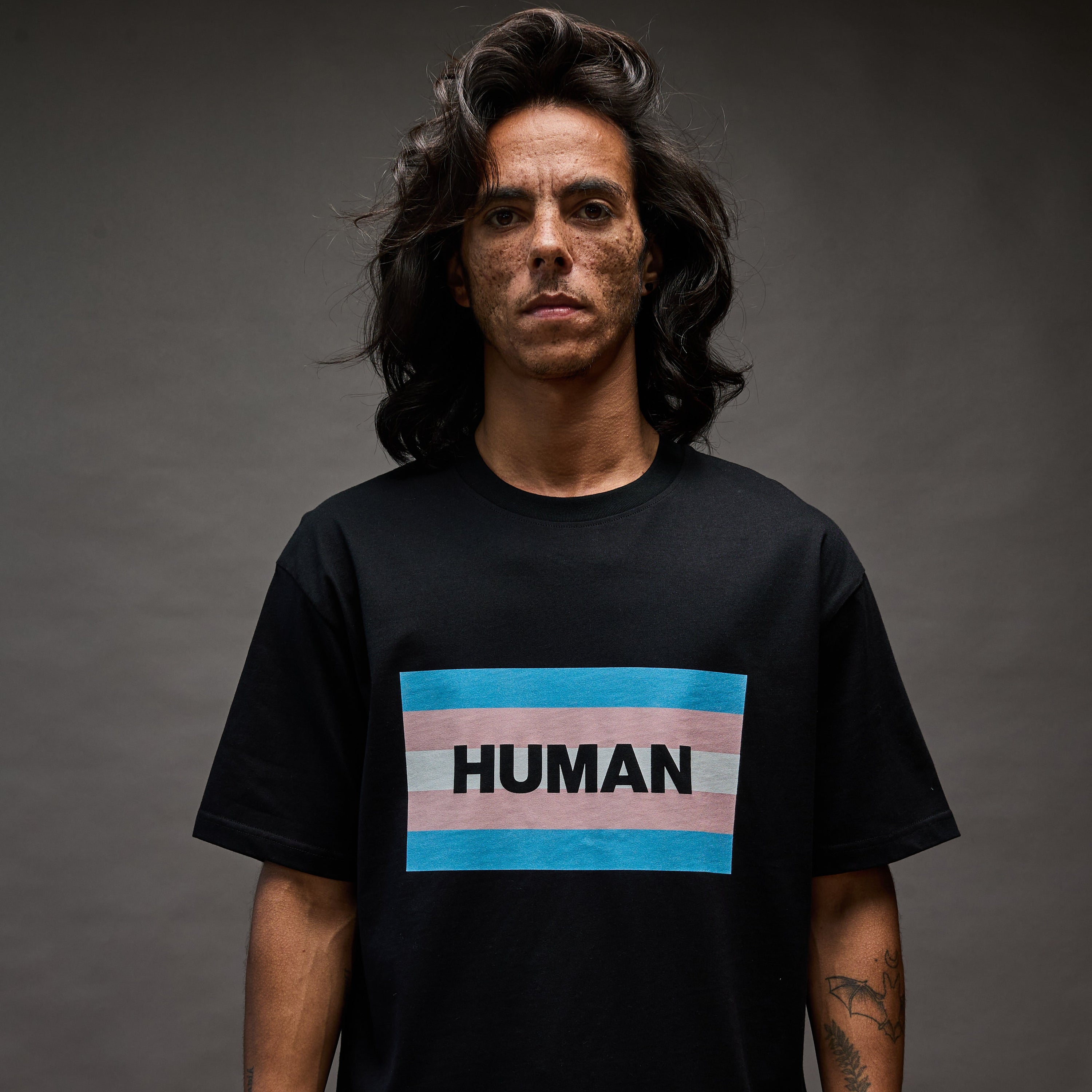 Person wearing a black t-shirt with a 'HUMAN' trans rights flag design on a dark background
